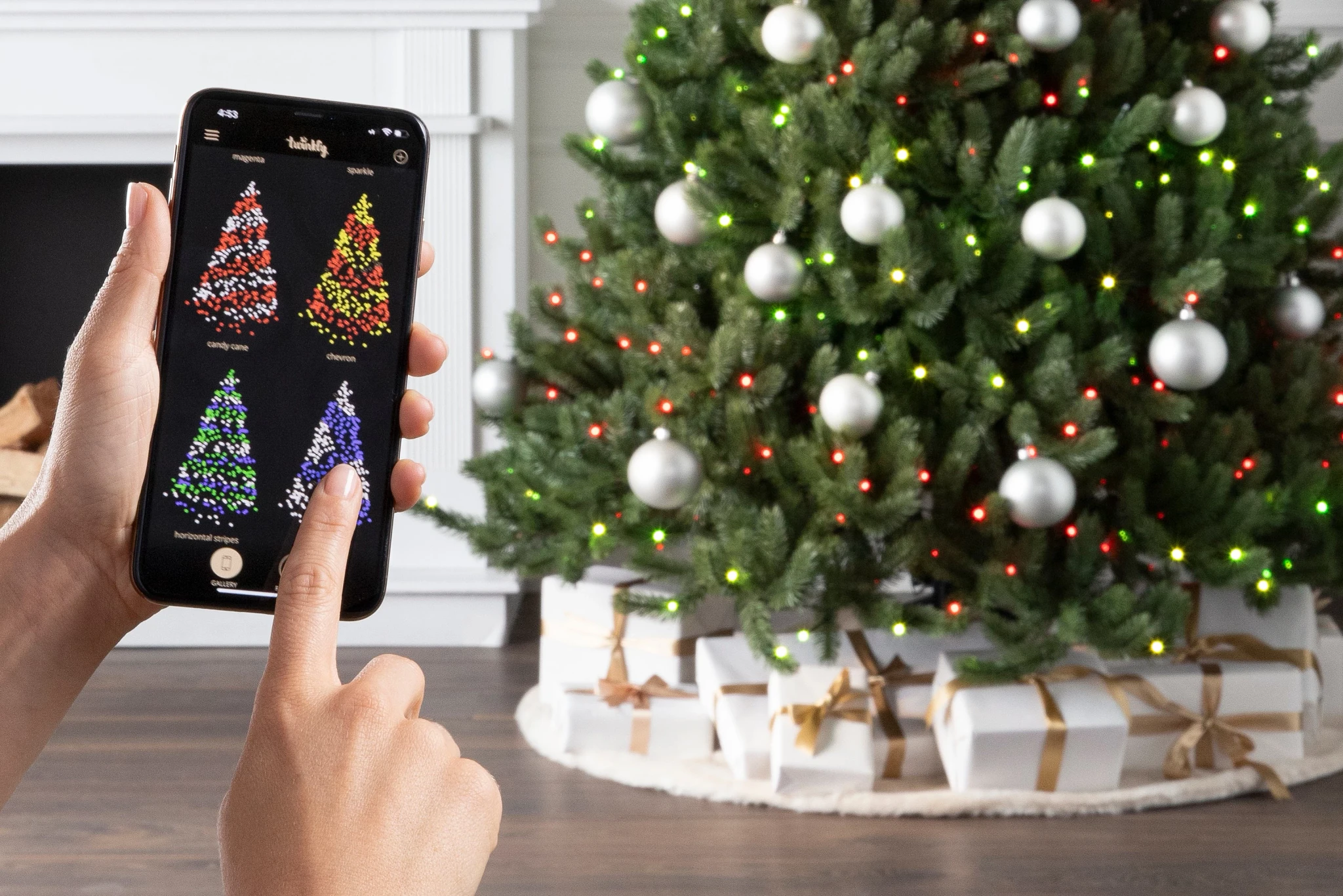 Person’s hands holding phone showing different types of lit Christmas trees with decorated Christmas tree in background