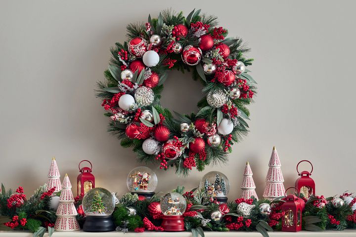 Mantel decorated with evergreen wreath with red and silver ornaments, snow globes, lanterns, and ceramic Christmas trees