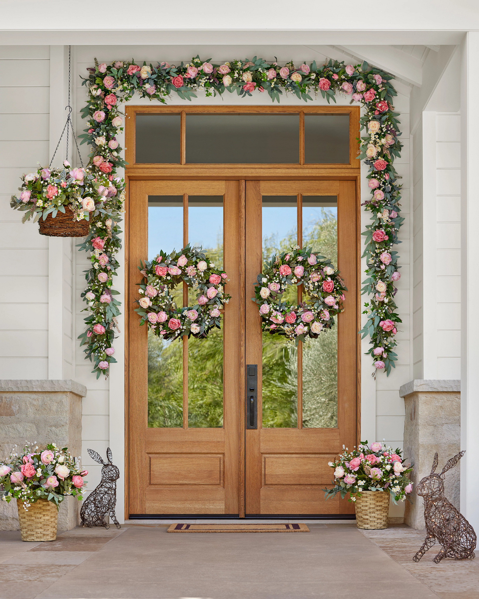 Brown double doors decorated with artificial flower wreaths, garlands, hanging baskets, and potted arrangements with faux cottage roses in various shades of pink