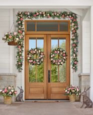 Brown double doors decorated with artificial flower wreaths, garlands, hanging baskets, and potted arrangements with faux cottage roses in various shades of pink