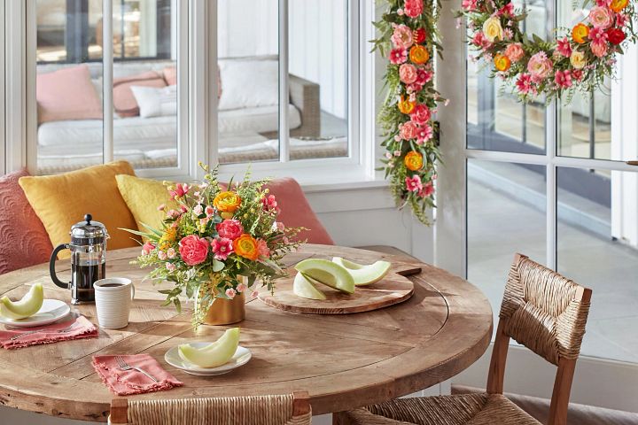 Round wooden dining table with artificial flower arrangement, a cup of coffee, a French press, and slices of honeydew on plates