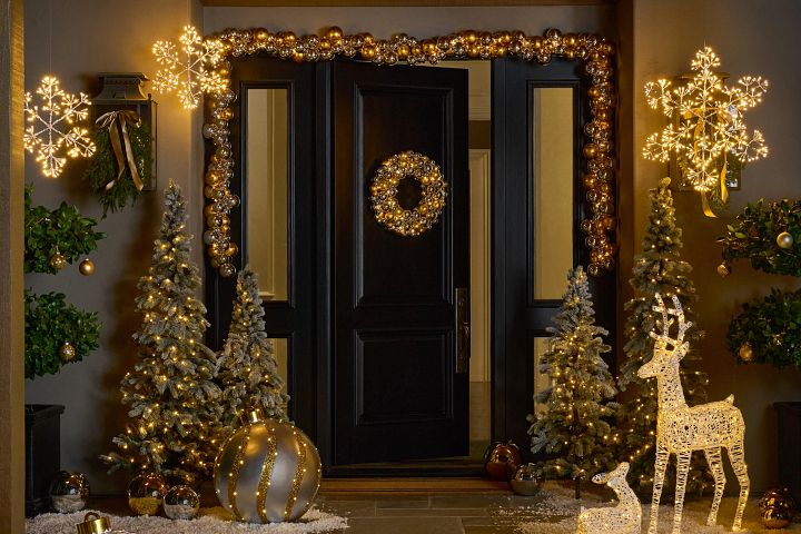 Entryway and black front door decorated with metallic wreath and garland, artificial potted trees, and lit deer wire sculptures