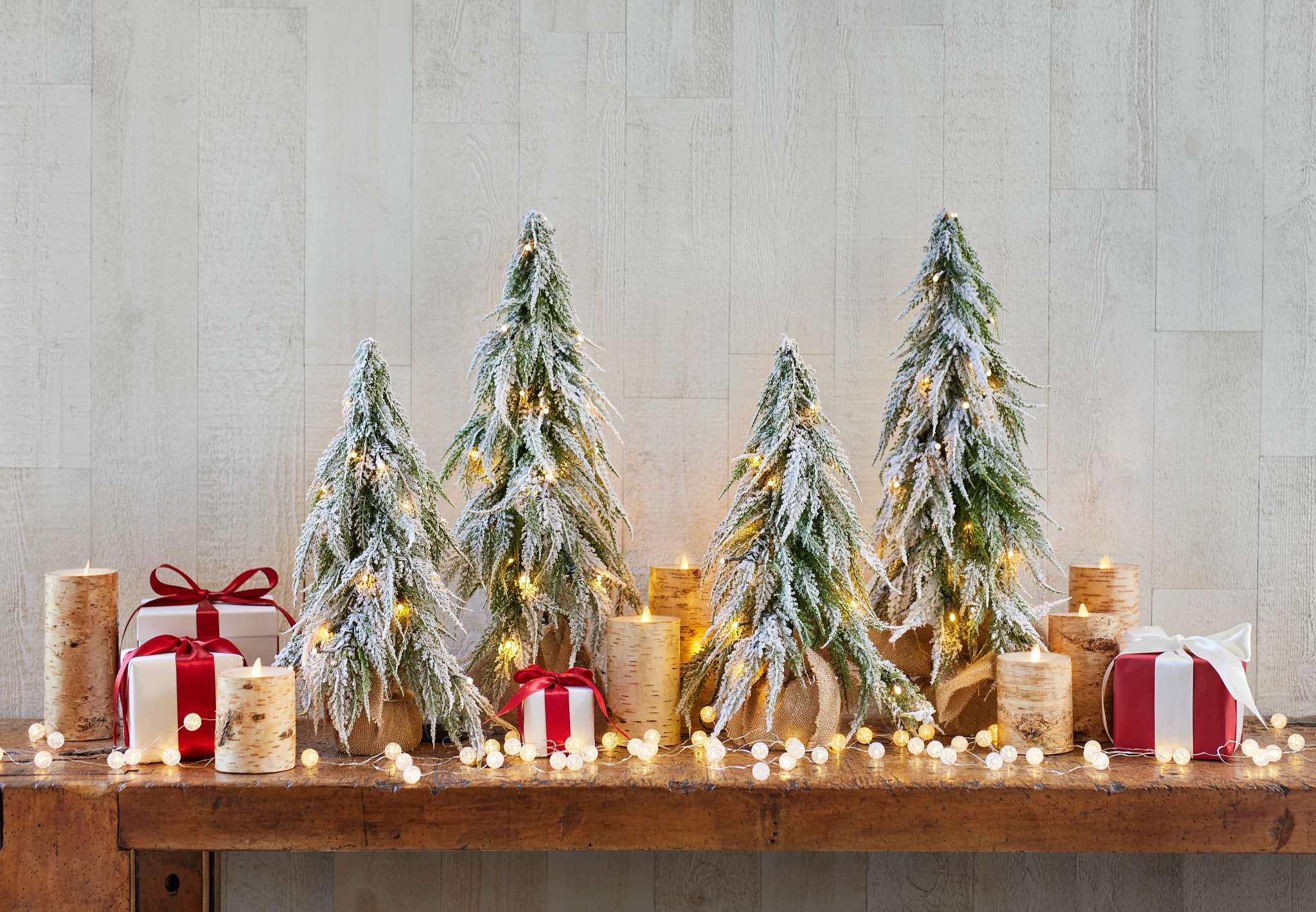 Frosted mini Christmas trees on a wooden table with rustic candles and wrapped presents