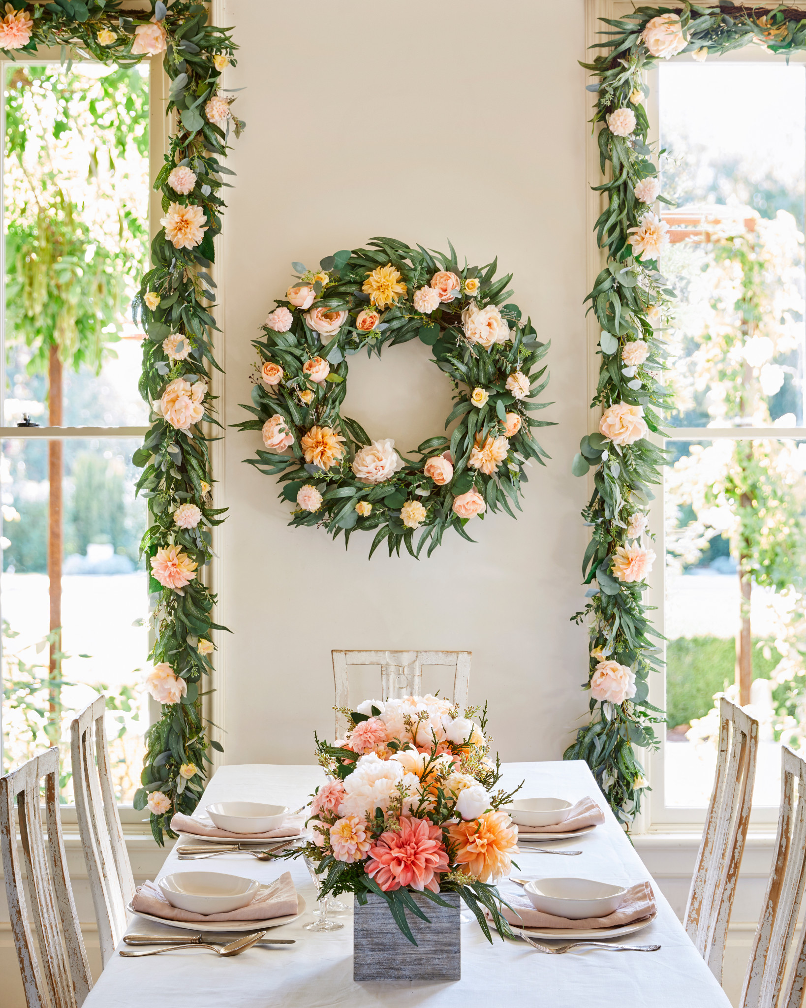 Indoor dining table decorated with artificial spring wreath, garland, and floral centerpiece featuring peonies, dahlias, roses, berries with buxus, eucalyptus, olive, and money leaves