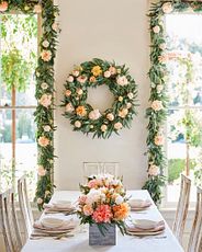 Indoor dining table decorated with artificial spring wreath, garland, and floral centerpiece featuring peonies, dahlias, roses, berries with buxus, eucalyptus, olive, and money leaves