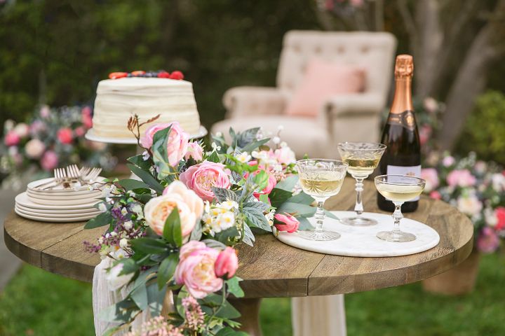 Outdoor dining table with artificial rose garland, cake, champagne bottle and glasses, and serveware on wooden table