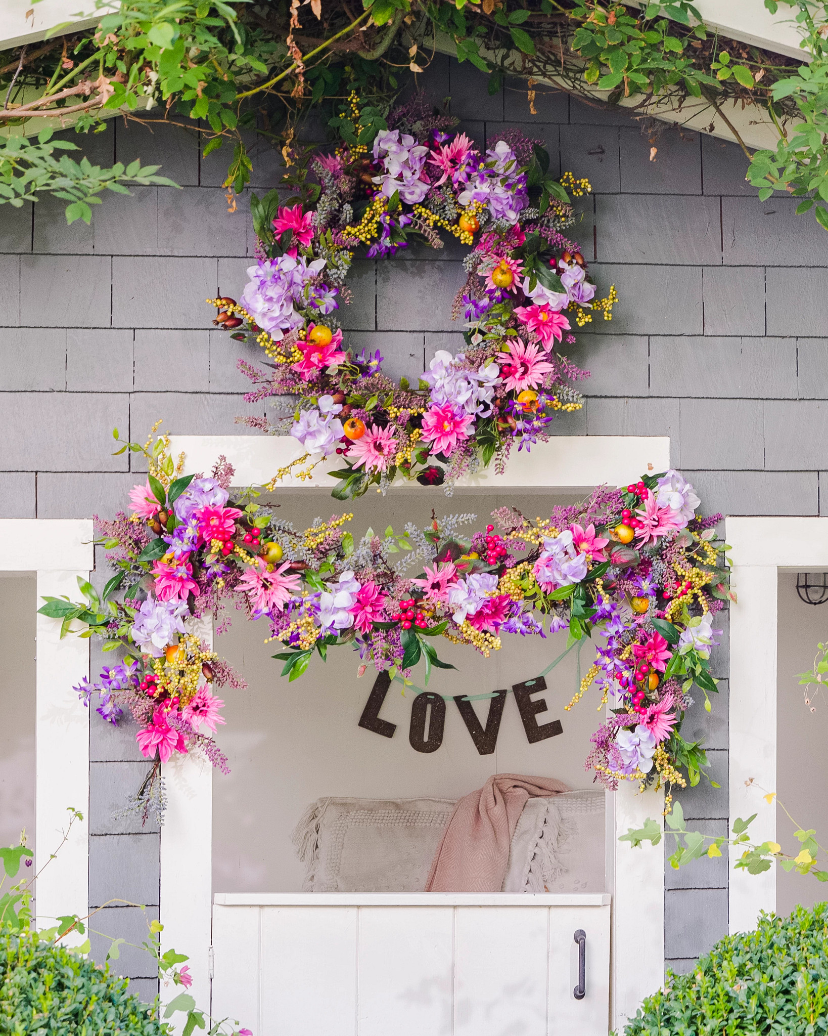 Facade of shed decorated with artificial spring wreath and garland featuring lavender, daisies, pomegranates, rose hips, purple hydrangeas, pink gerberas, mixed berries, and leaves