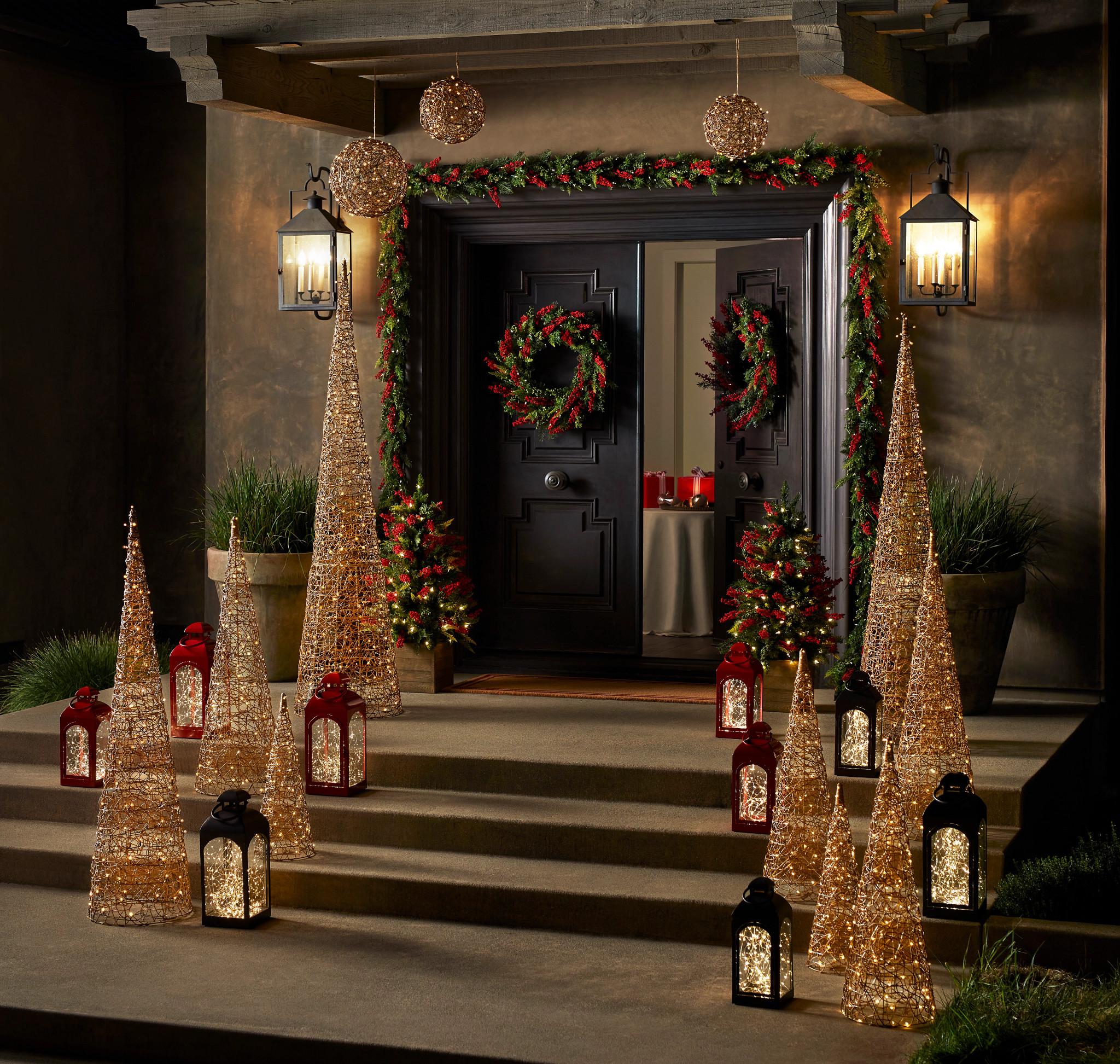 Black double doors decorated with artificial Christmas wreath and garland and front steps with lit cone wire trees and red and black lanterns with fairy lights