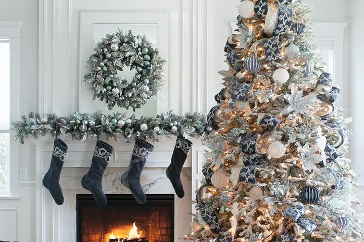 Decorated frosted Christmas tree next to fireplace decorated with frosted wreath, garland, and blue velvet stockings