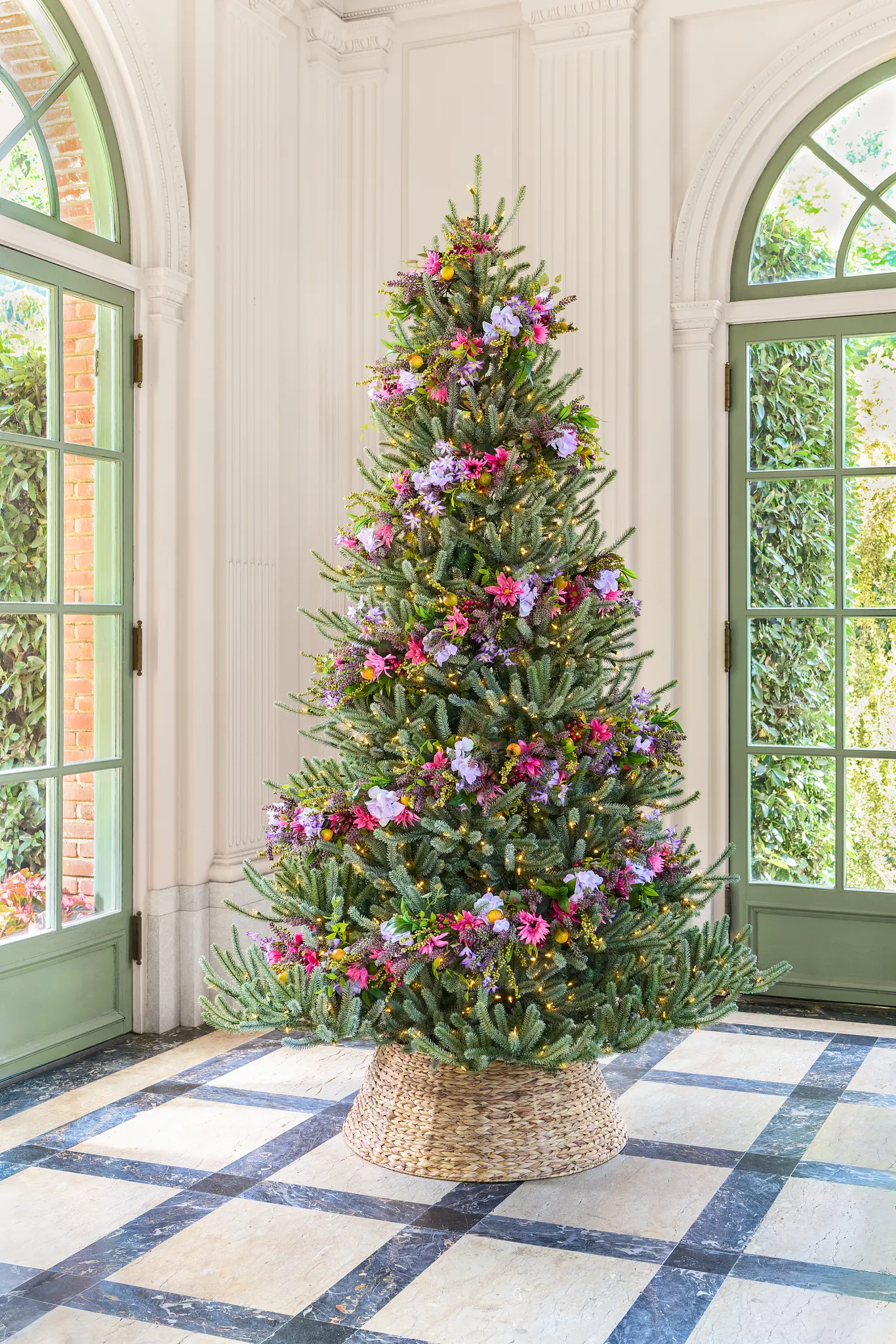 A beautifully decorated Christmas tree adorned with colorful flowers and lights, set in a bright room with large green windows.