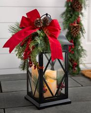 Black lantern with three candles decorated with pinecones, red wired bow, bells, red berries, and mixed foliage